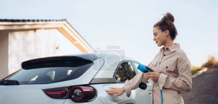 Junge Frau hält Stromkabel aus ihrem Auto, bereit für das Laden zu Hause, nachhaltiges und wirtschaftliches Transportkonzept.