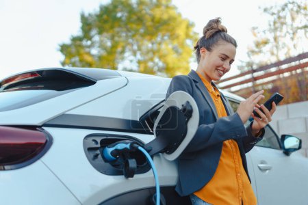Mujer joven con smartphone esperando mientras se carga el coche eléctrico en la estación de carga del hogar, concepto de transporte sostenible y económico.