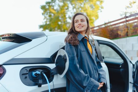 Mujer joven esperando mientras se carga el coche eléctrico en la estación de carga en el hogar, concepto de transporte sostenible y económico.