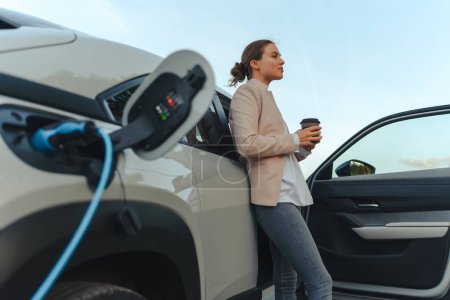 Young woman with cup of coffee waiting while electric car charging, sustainable and economic transportation concept.