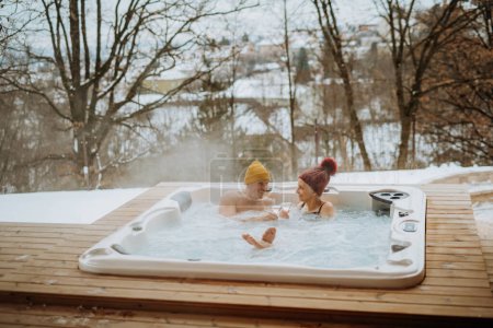 Téléchargez les photos : Couple sénior en bonnet tricoté profitant ensemble d'une baignoire extérieure avec verre de vin sur leur terrasse lors d'une froide journée d'hiver. - en image libre de droit