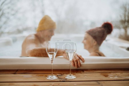 Photo for Senior couple in knitted cap enjoying together outdoor bathtub with glass of wine at their terrace during a cold winter day. - Royalty Free Image