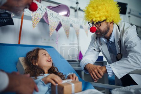 Photo for Happy doctor with clown red noses celebrating birthday with little girl in a hospital room. - Royalty Free Image