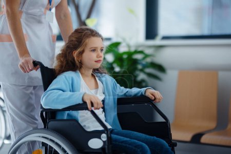 Krankenschwester schubst kleines Mädchen im Rollstuhl auf Krankenhausflur.