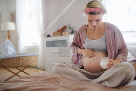 Foto de Mujer embarazada haciendo crujir su vientre, sentada en una cama, cuidando de sí misma. - Imagen libre de derechos