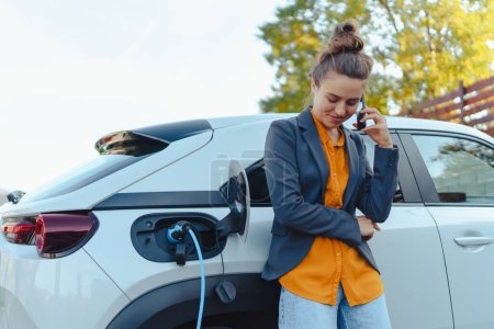 Mujer joven con smartphone esperando mientras se carga el coche eléctrico en la estación de carga del hogar, concepto de transporte sostenible y económico.