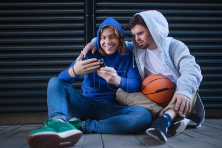 Photo for Man with down syndrome resting during basketball playing outdoor with his friend. Concept of friendship and integration people with disability into a society. - Royalty Free Image