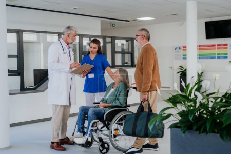 Foto de Médico maduro y su joven colega conversando con sus pacientes mayores en el pasillo del hospital. - Imagen libre de derechos