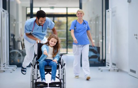 Photo for Young doctor pushing little girl at wheelchair at the pediatric corridor, having fun. - Royalty Free Image