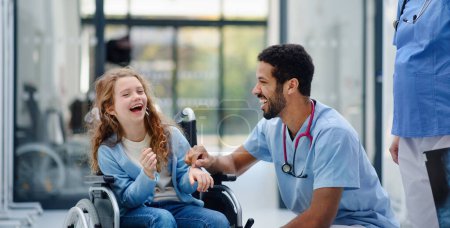 Téléchargez les photos : Jeune médecin multiracial s'amuser avec une petite fille sur un fauteuil roulant. - en image libre de droit