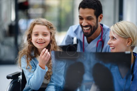 Téléchargez les photos : Jeune médecin multiracial avec son collègue montrant une image radiographique des poumons à un petit patient en fauteuil roulant. - en image libre de droit