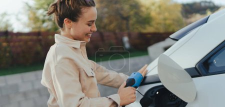 Young woman holding power supply cable from her car, prepared for charging it in home, sustainable and economic transportation concept.