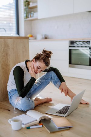 Téléchargez les photos : Jeune femme assise avec ordinateur portable et tasse de café dans sa cuisine. - en image libre de droit