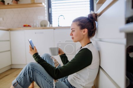 Foto de Mujer joven sentada con teléfono inteligente y taza de café en su cocina. - Imagen libre de derechos