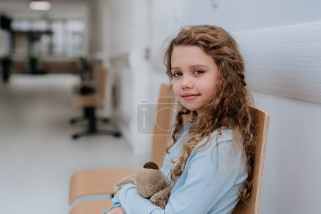 Téléchargez les photos : Portrait de petite fille avec un ours en peluche assis et attendant dans le couloir de l'hôpital. - en image libre de droit