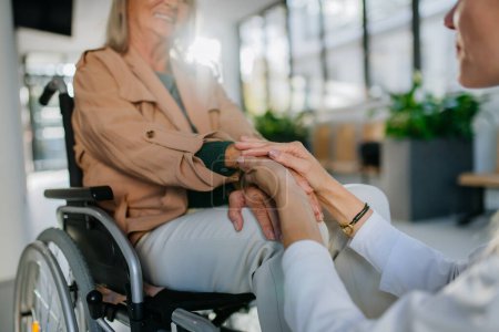 Téléchargez les photos : Gros plan d'un jeune médecin tenant la main d'une patiente en fauteuil roulant, la consolant. - en image libre de droit