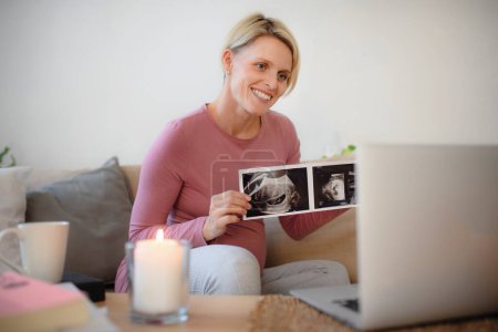 Photo for Pregnant woman showing ultrasound photo of a baby to webcam. - Royalty Free Image