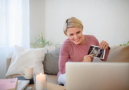 Téléchargez les photos : Femme enceinte montrant la photo échographique d'un bébé à webcam. - en image libre de droit