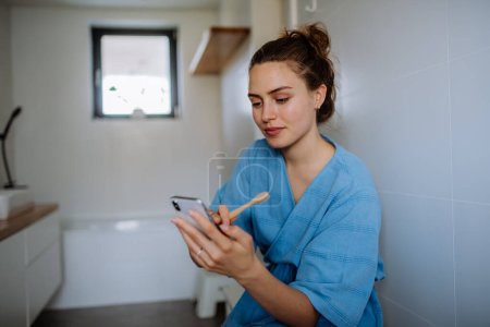 Photo for Young woman brushing teeth and scrolling her smartphone, morning routine concept. - Royalty Free Image
