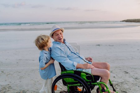 Téléchargez les photos : Petit garçon poussant son grand-père sur le fauteuil roulant, profitant de la mer ensemble. - en image libre de droit