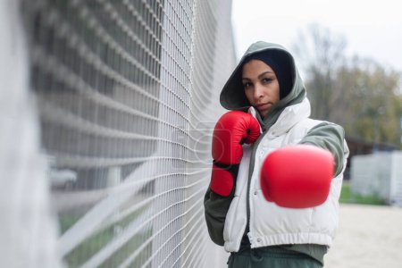 Téléchargez les photos : Portrait d'une jeune femme musulmane avec des gants de boxe debout en plein air dans une ville. - en image libre de droit