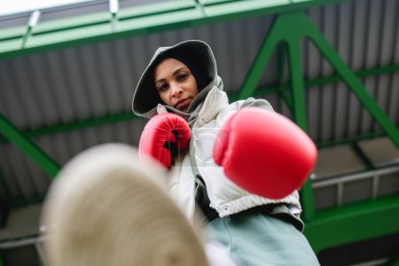 Téléchargez les photos : Vue en angle bas d'une jeune femme musulmane avec des gants de boxe debout en plein air dans une ville. - en image libre de droit
