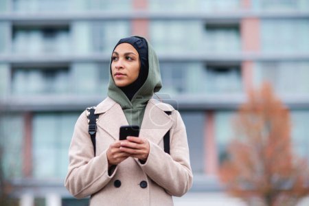 Téléchargez les photos : Portrait de jeune femme musulmane en manteau, debout en plein air dans une ville, smartphone défilant. - en image libre de droit