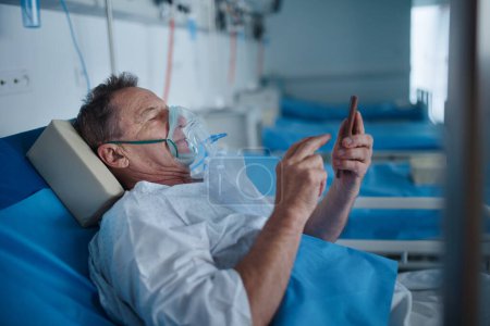 Photo for Senior man with oxygen mask lying in the hospital bed and scrolling smartphone. - Royalty Free Image