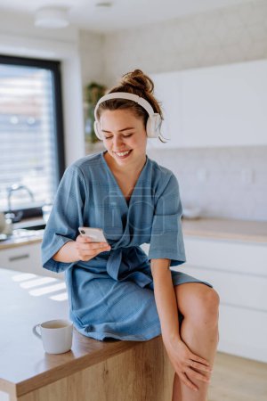 Foto de Mujer joven disfrutando de una taza de café, el teléfono inteligente de desplazamiento y escuchar música por la mañana, en una cocina. - Imagen libre de derechos