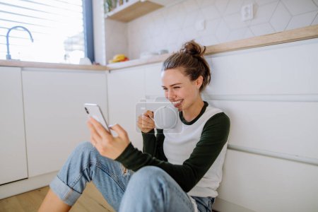 Téléchargez les photos : Jeune femme assise avec smartphone et tasse de café dans sa cuisine. - en image libre de droit