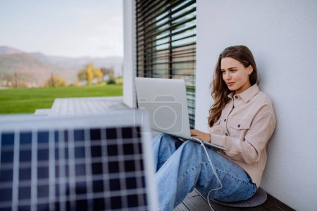 Téléchargez les photos : Jeune femme assise sur la terrasse, chargeant tablette à travers le panneau solaire. - en image libre de droit