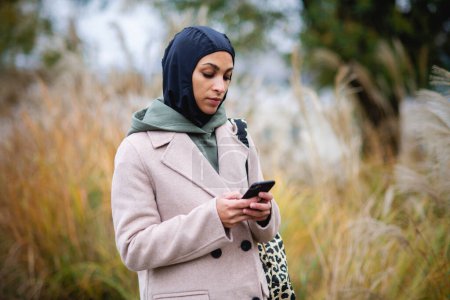 Téléchargez les photos : Portrait de jeune femme musulmane en manteau, debout en plein air dans un parc de la ville, smartphone défilant. - en image libre de droit