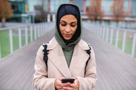 Téléchargez les photos : Portrait de jeune femme musulmane en manteau, debout en plein air dans une ville, smartphone défilant. - en image libre de droit