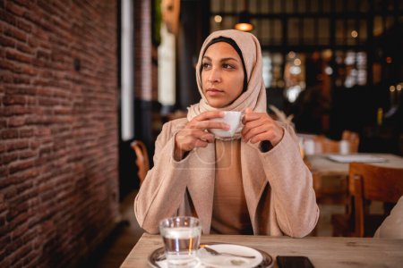 Téléchargez les photos : Jeune femme musulmane dégustant une tasse de café dans un café. - en image libre de droit