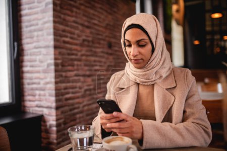 Foto de Mujer musulmana joven sentada en la cafetería y desplazando un teléfono inteligente. - Imagen libre de derechos