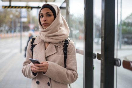 Photo for Young muslim woman with a smartphone waiting for bus at city bus stop. - Royalty Free Image