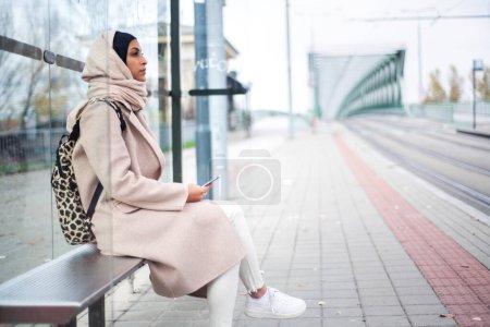 Photo for Young muslim woman waiting for a bus at city bus stop. - Royalty Free Image