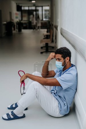 Foto de Joven médico angustiado sentado en el piso de un pasillo del hospital. - Imagen libre de derechos