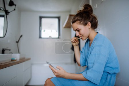 Foto de Mujer joven cepillarse los dientes y desplazar su teléfono inteligente, concepto de rutina de la mañana. - Imagen libre de derechos