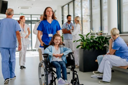 Téléchargez les photos : Jeune infirmière poussant une petite fille en fauteuil roulant dans un couloir d'hôpital. - en image libre de droit