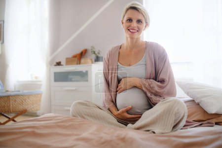Photo for Happy pregnant woman stroking her belly sitting on a bed. - Royalty Free Image