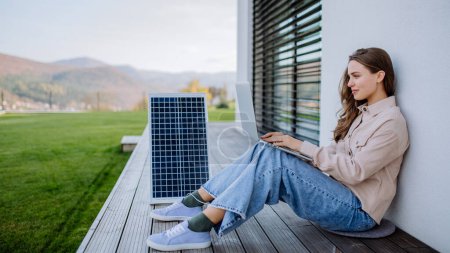 Téléchargez les photos : Jeune femme assise sur la terrasse, chargeant tablette à travers le panneau solaire. - en image libre de droit