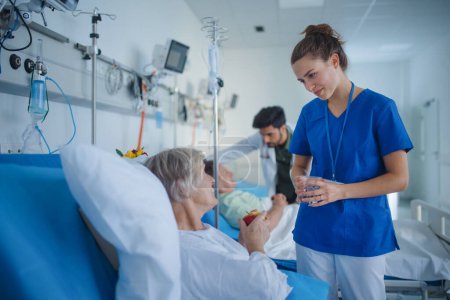Téléchargez les photos : Jeune infirmière donnant un verre d'eau à un patient âgé dans un service hospitalier. - en image libre de droit