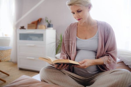 Foto de Mujer embarazada sentada en la cama y leyendo un libro. - Imagen libre de derechos
