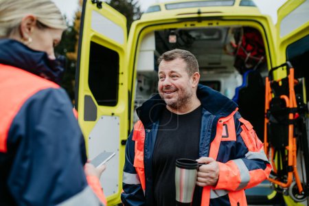 Photo for Rescuers having break in front of an ambulance car, talking and drinking coffee. - Royalty Free Image
