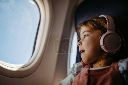 Photo for Little girl in airplane listening music and looking out of the window. - Royalty Free Image