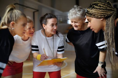 Téléchargez les photos : Un entraîneur de sport féminin avec presse-papiers discutant de tactiques avec des jeunes et des vieilles femmes entraînement par équipe pour le match dans la salle de gym. - en image libre de droit