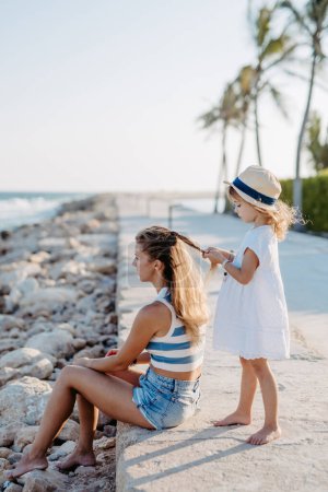 Téléchargez les photos : Little girl making hairstyle her mother, enjoying summer time at ocean. - en image libre de droit