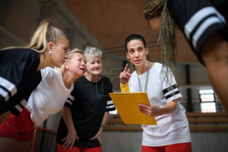 Téléchargez les photos : Un entraîneur de sport féminin avec presse-papiers discutant de tactiques avec des jeunes et des vieilles femmes entraînement par équipe pour le match dans la salle de gym. - en image libre de droit