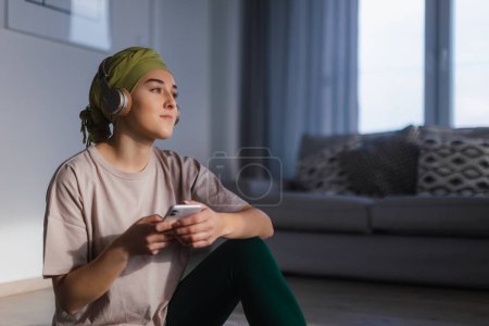 Foto de Mujer joven con cáncer sentada en casa y desplazando un teléfono y escuchando la música. - Imagen libre de derechos